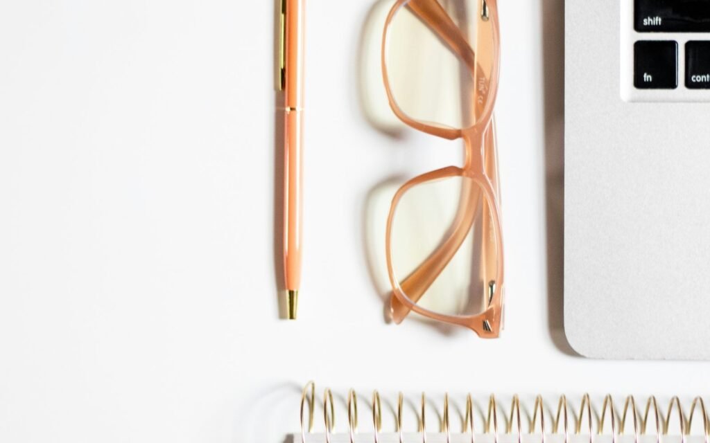A minimal white workspace with a notebook, pen, glasses and laptop.